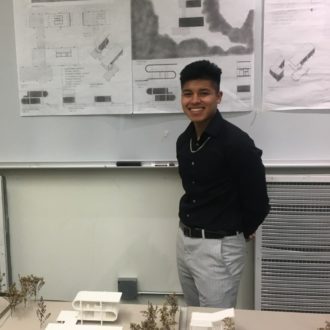 A male ������ student poses in front of a poster with a building design while a 3-D model of the building rests on a table in front of him